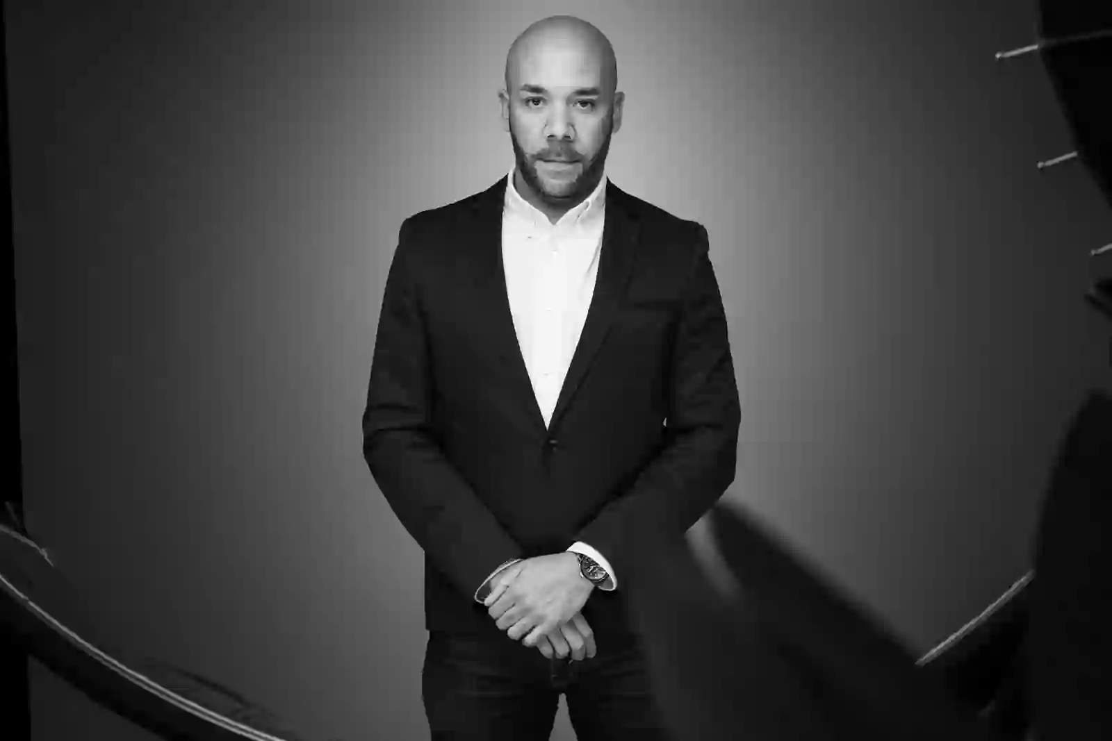 Portrait en noir et blanc d’un homme à barbe courte en veste sombre, les mains croisées devant lui