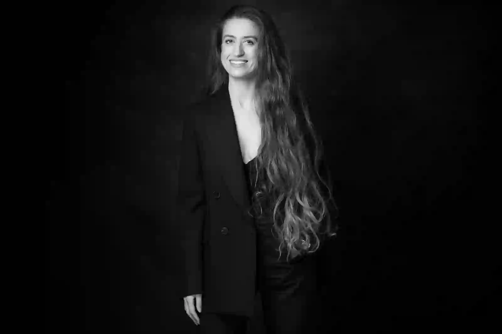 Portrait en noir et blanc d’une femme souriante aux très longs cheveux ondulés, en tenue sombre