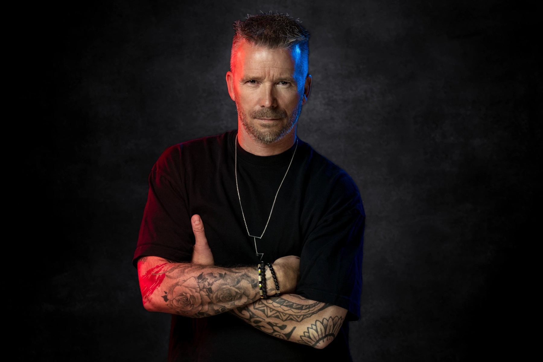 Homme en tee-shirt noir, bras tatoués croisés, sous un éclairage rouge et bleu