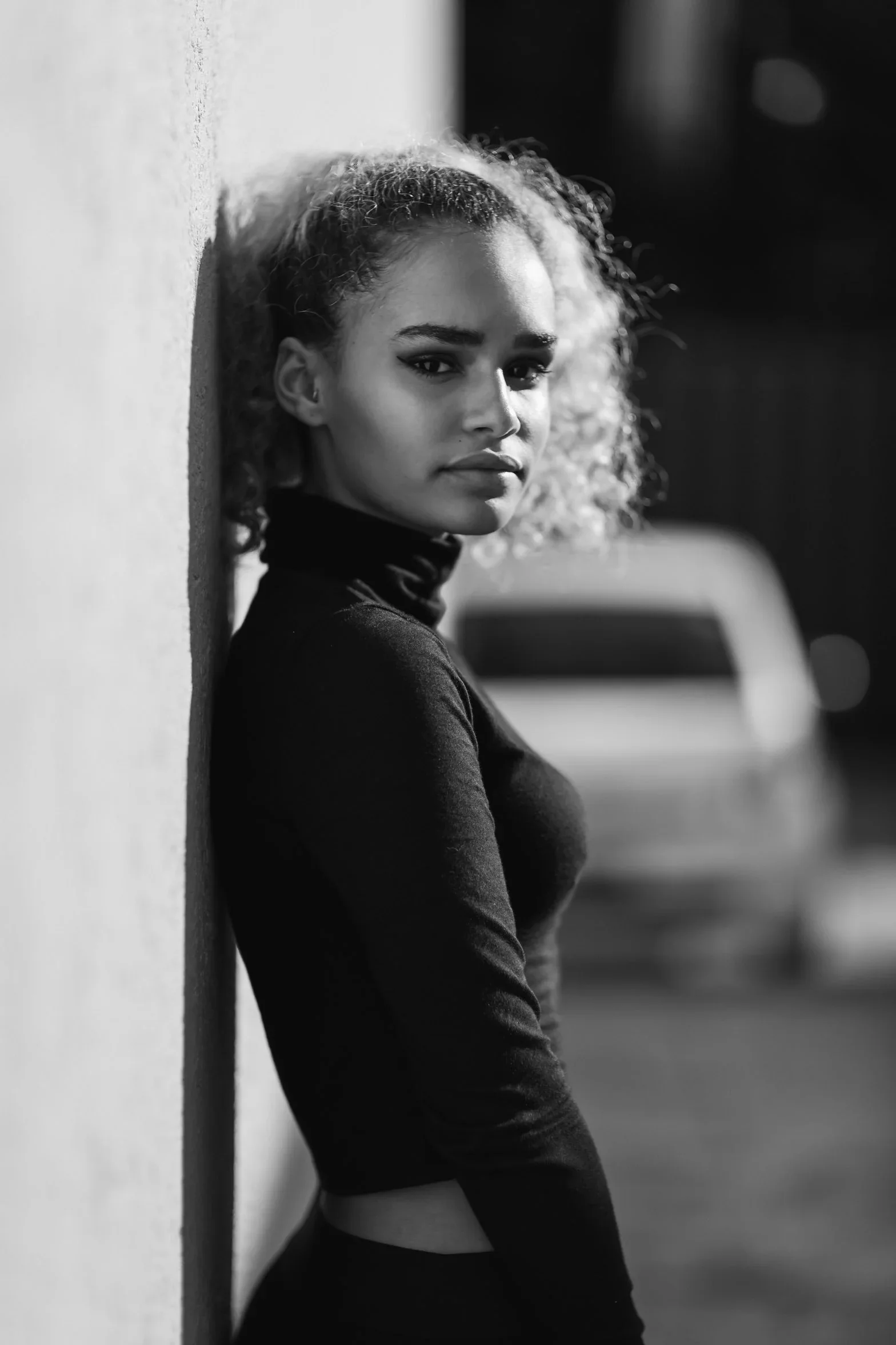 Portrait en noir et blanc d’une femme en col roulé appuyée contre un mur clair