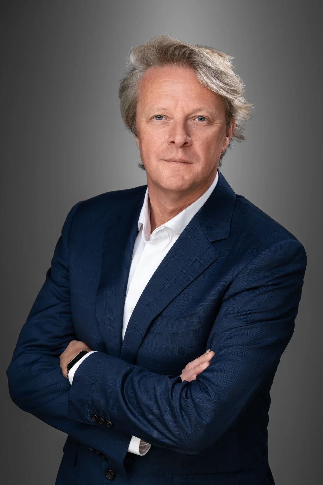 Homme aux cheveux gris, bras croisés, en veste bleu foncé et chemise blanche, sur fond gris